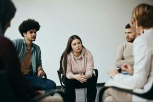 A group sits in a circle engaged in group counseling with a licensed medical professional guiding the session.