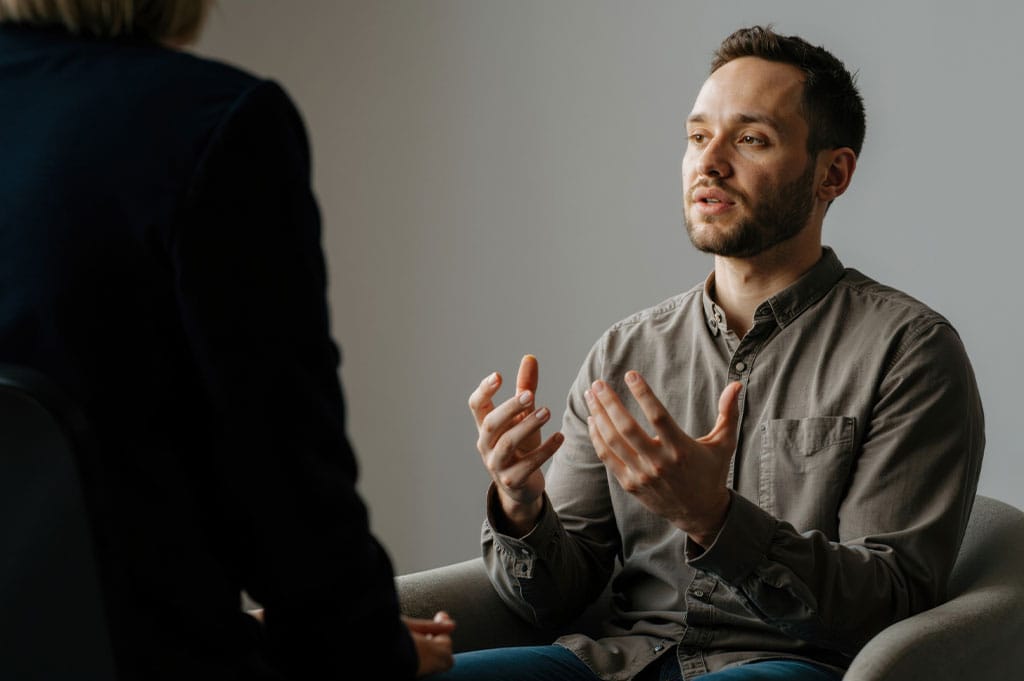 A man in a button-down shirt speaks openly during a therapy session, gesturing as he explains something to a clinician.