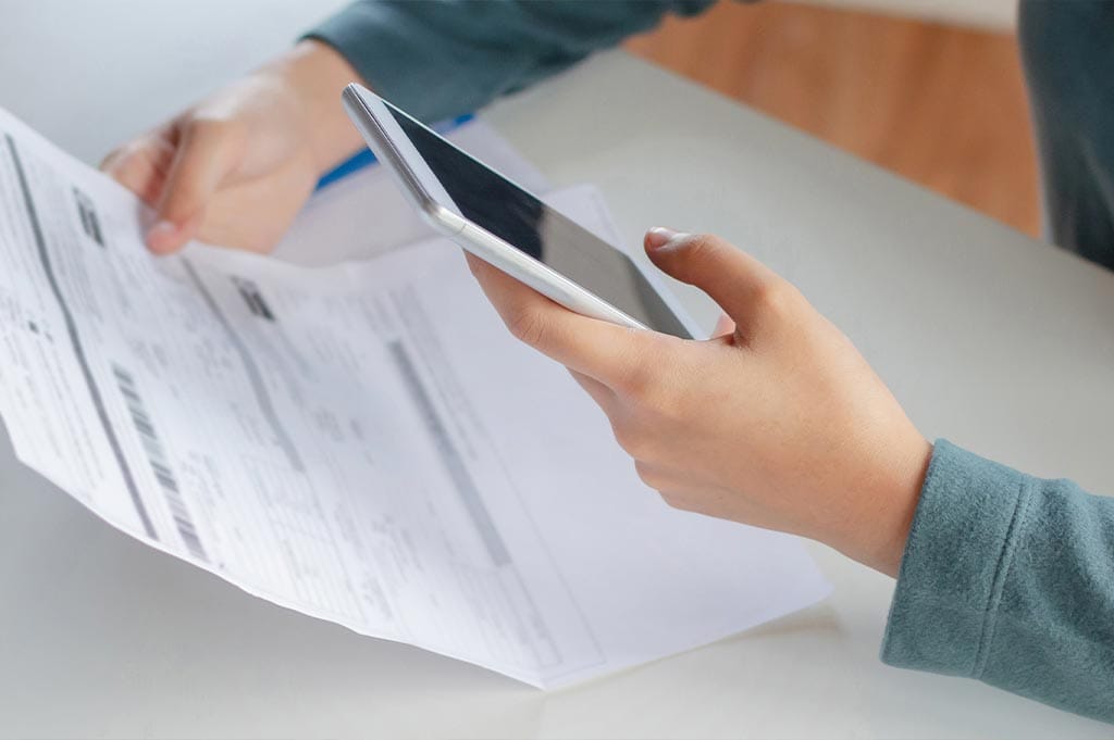 woman sitting at a table with her phone reviewing her insurance policy to see how much alcohol rehab would cost