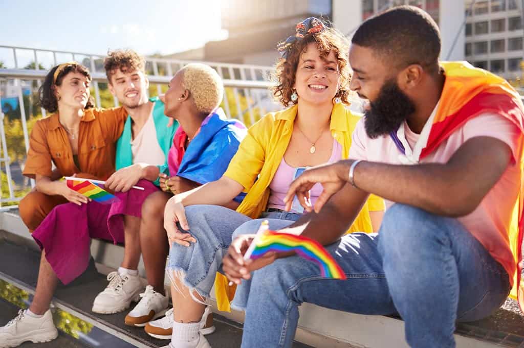LGBTQ+ individuals sitting together outdoors holding Pride flags, representing community, connection, and belonging in recovery